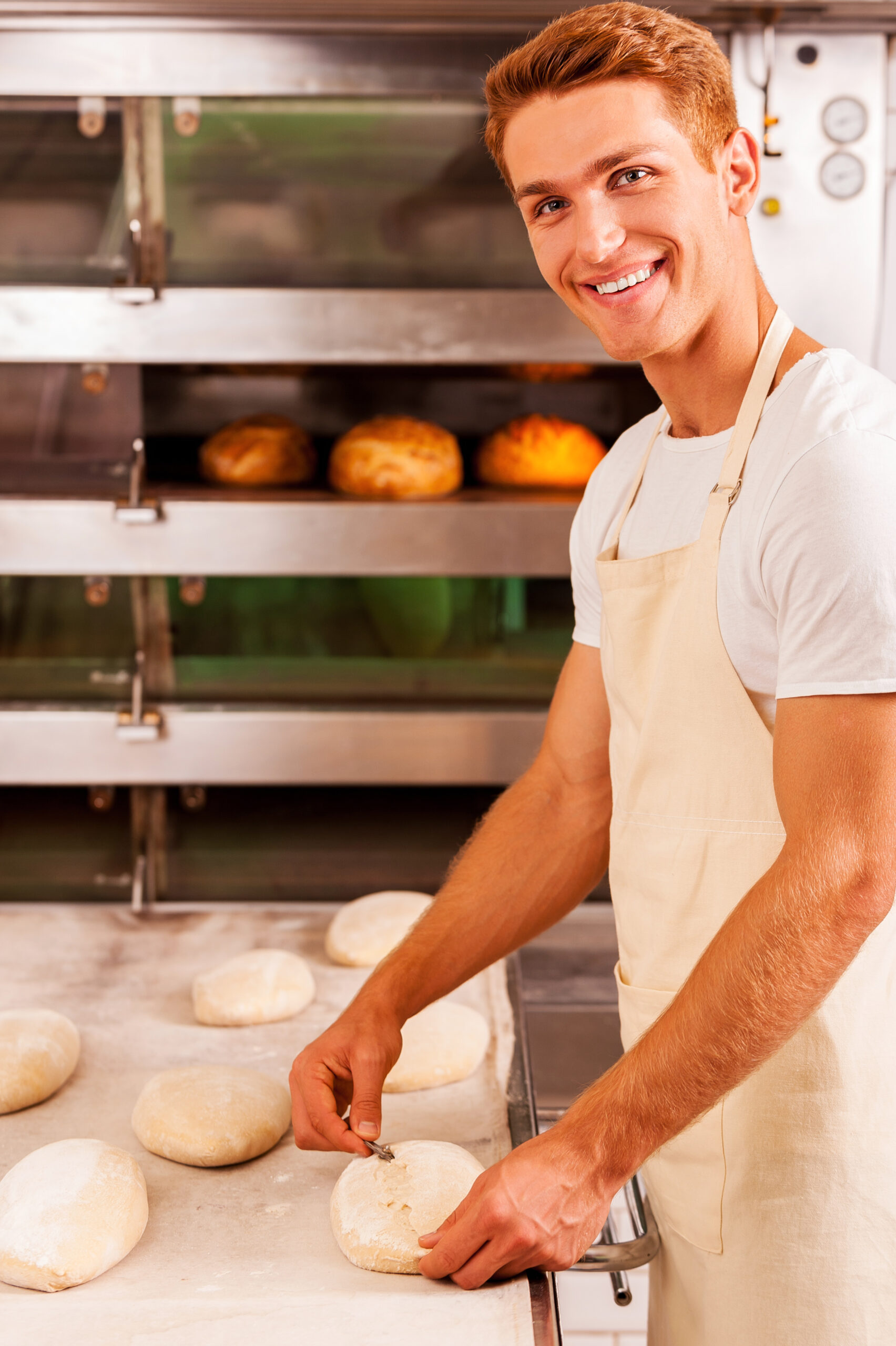 Confident baker at work. Confident young man in apron working with dough and smiling while standing against oven with bread in it